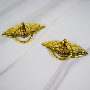 May include: Two gold-toned metal drawer pulls with ornate designs and ring handles. The pulls are on a white marble surface.
