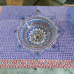 Op de afbeelding: Een blauwe en witte keramische wastafel met een geometrisch patroon, ingebouwd in een aanrechtblad met een blauw en wit moza&iuml;ek tegelontwerp. Het aanrechtblad heeft een rode en witte rand met een bloemmotief.