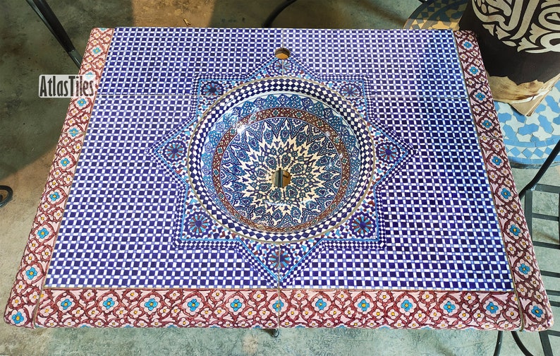 Op de afbeelding: Een blauwe en witte keramische wastafel met een geometrisch patroon, ingebouwd in een aanrechtblad met een blauw en wit moza&iuml;ek tegelontwerp. Het aanrechtblad heeft een rode en witte rand met een bloemmotief.
