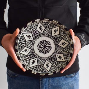 May include: A large, round, black and white ceramic plate with a geometric pattern. The plate has a raised rim and a center design that resembles a star.