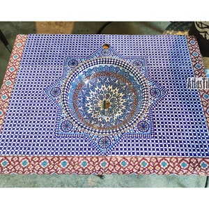 Op de afbeelding: Een blauwe en witte keramische wastafel met een geometrisch patroon, ingebouwd in een aanrechtblad met een roze en blauwe rand. De wastafel is omgeven door een decoratief stervormig ontwerp. Het aanrechtblad is gemaakt van keramische tegels.
