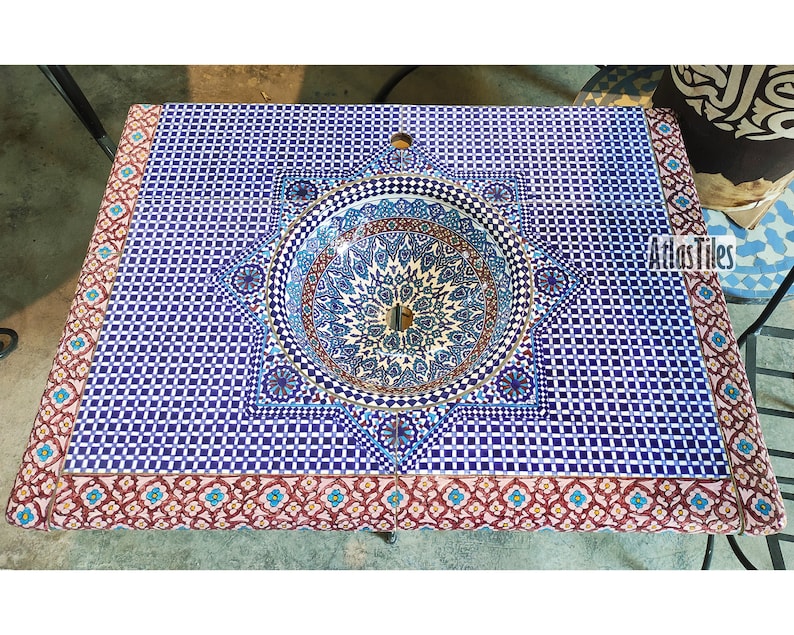 Op de afbeelding: Een blauwe en witte keramische wastafel met een geometrisch patroon, ingebouwd in een aanrechtblad met een roze en blauwe rand. De wastafel is omgeven door een decoratief stervormig ontwerp. Het aanrechtblad is gemaakt van keramische tegels.