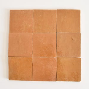 以下が含まれることがあります： 3x3のグリッドに配置された9枚のテラコッタタイル。タイルは暖かみのある赤褐色で、表面がわずかに不均一です。