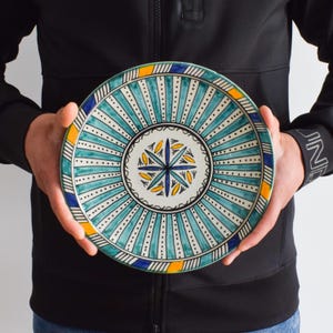 May include: A large, round ceramic plate with a blue, white, and yellow geometric design. The plate has a central starburst pattern surrounded by concentric circles of stripes and dots.