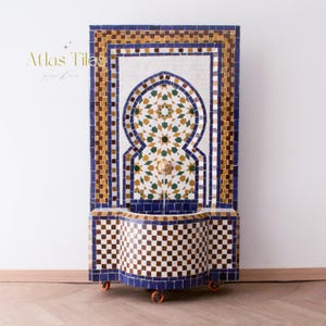 May include: A mosaic tile fountain with a Moorish design. The fountain features a central arch with a geometric pattern in green, gold, and white. The frame is blue, brown, and white. The base has a checkered pattern. The text "Atlas Tilas" is in the upper left corner.