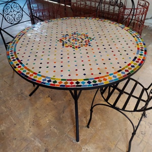 Op de afbeelding: Ronde mozaïektafel met een witte basis en een kleurrijk mozaïekblad. De tafel heeft een zwart metalen frame en poten. Het mozaïekblad heeft een geometrisch patroon met verschillende kleuren, waaronder blauw, groen, rood, geel en oranje.