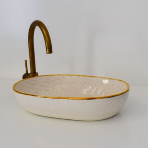 Pode incluir: Pia de cerâmica oval branca com borda dourada e torneira de latão.