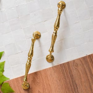 Puede incluir: Dos tiradores de puerta dorados con diseños intrincados. Los tiradores son de metal y tienen un acabado pulido.