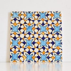 May include: A square tile with a geometric pattern of blue, orange, and white stars on a white background.