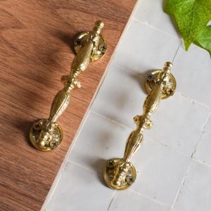 Puede incluir: Dos tiradores de cajón de metal dorado con diseños ornamentados. Los tiradores tienen una base redondeada y un mango largo y delgado.