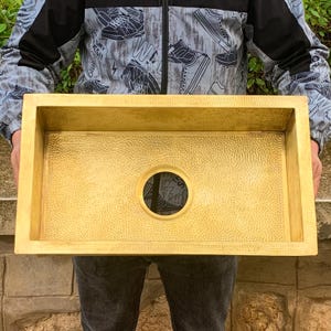 Könnte beinhalten: Ein großes, rechteckiges, gehämmertes Küchenspülbecken aus Messing mit einem Abflussloch. Das Spülbecken ist hellgoldfarben und hat eine strukturierte Oberfläche.