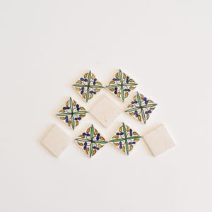 以下が含まれることがあります： 白い背景に青、緑、黄色の花柄が描かれた装飾的なセラミックタイル9枚。タイルは白い表面にピラミッド型に配置されています。