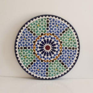 May include: A round, decorative tray with a Moroccan-inspired geometric pattern in shades of blue, green, yellow, and brown. The tray features a central starburst design.