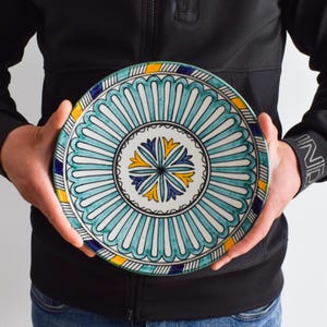 May include: A large, round ceramic plate with a blue, green, and yellow geometric design. The plate has a white center with a blue and yellow floral pattern.