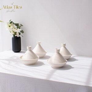 May include: Four white ceramic tagines with lids, arranged on a white tablecloth. The tagines are a traditional Moroccan cookware.