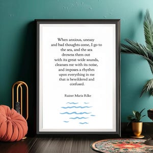 May include: A framed print with a black border features a poem by Rainer Maria Rilke. The text is in black on a white background, with blue wave illustrations. A coral-coloured decorative cushion and a gold-coloured decorative object are in the foreground.