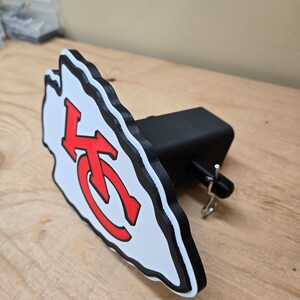 K&ouml;nnte beinhalten: Ein wei&szlig;es und rotes Logo der Kansas City Chiefs, das an einem schwarzen Anh&auml;ngerkupplungsschutz befestigt ist. Das Logo ist ein stilisierter Pfeilspitz mit den Buchstaben "KC" darin.