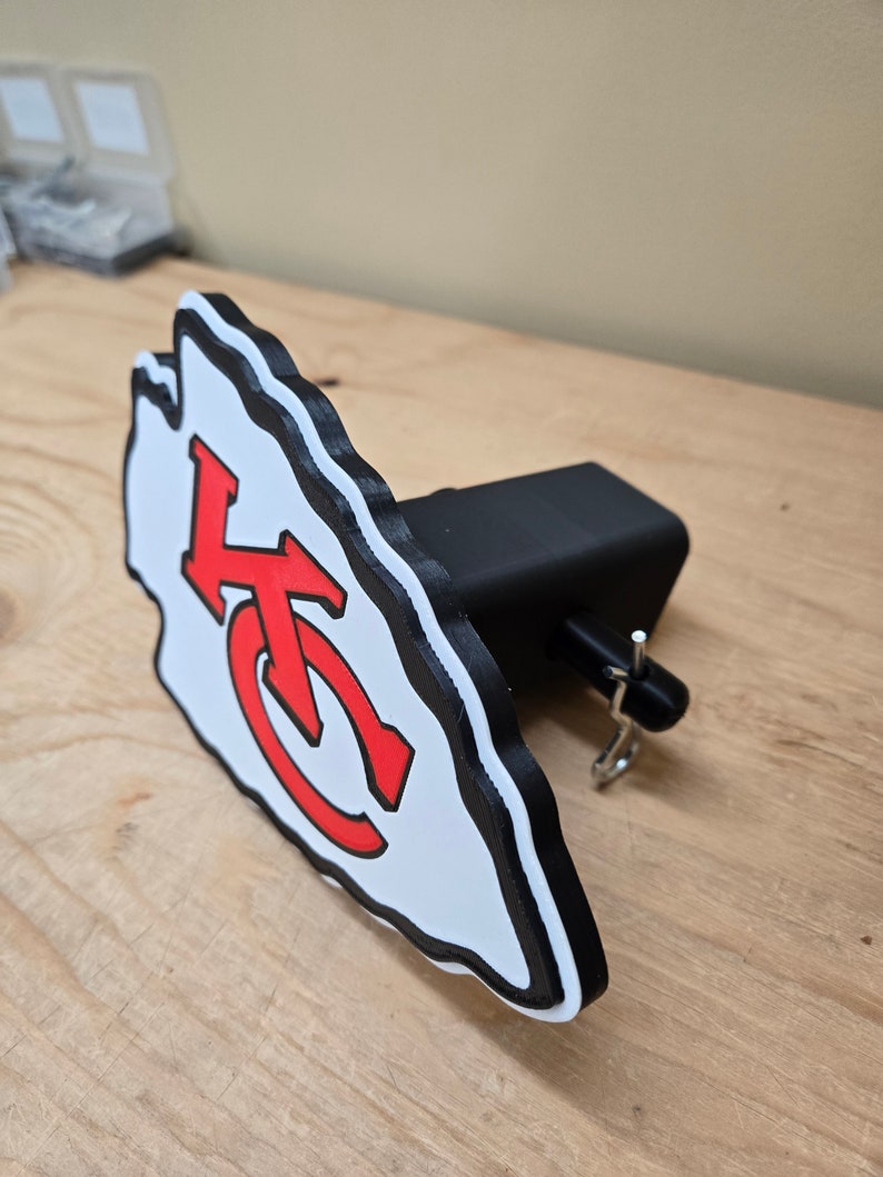 K&ouml;nnte beinhalten: Ein wei&szlig;es und rotes Logo der Kansas City Chiefs, das an einem schwarzen Anh&auml;ngerkupplungsschutz befestigt ist. Das Logo ist ein stilisierter Pfeilspitz mit den Buchstaben "KC" darin.