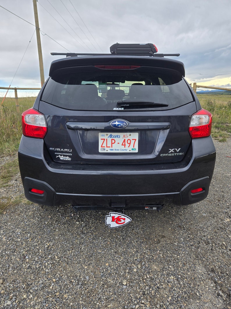K&ouml;nnte beinhalten: Ein schwarzer Subaru XV Crosstrek mit einem Nummernschild "ZLP-497" und einem Kansas City Chiefs-Logo an der Anh&auml;ngerkupplung. Das Auto hat ein Dachgep&auml;cktr&auml;ger mit einer schwarzen Box auf dem Dach.