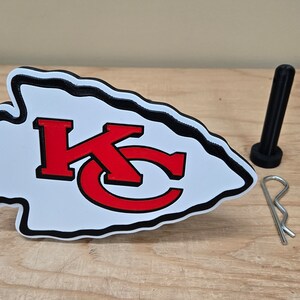 K&ouml;nnte beinhalten: Ein schwarzes pfeilf&ouml;rmiges Emblem mit einem wei&szlig;en und roten Logo der Kansas City Chiefs. Das Emblem ist aus Kunststoff und hat einen schwarzen Rand. Das Logo ist ein stilisiertes "KC" in Rot mit schwarzem Umriss. Das Emblem befindet sich auf einer Holzoberfl&auml;che mit einem schwarzen Plastik-Stift und einer silbernen Sicherheitsnadel.