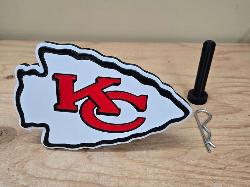 K&ouml;nnte beinhalten: Ein schwarzes pfeilf&ouml;rmiges Emblem mit einem wei&szlig;en und roten Logo der Kansas City Chiefs. Das Emblem ist aus Kunststoff und hat einen schwarzen Rand. Das Logo ist ein stilisiertes "KC" in Rot mit schwarzem Umriss. Das Emblem befindet sich auf einer Holzoberfl&auml;che mit einem schwarzen Plastik-Stift und einer silbernen Sicherheitsnadel.