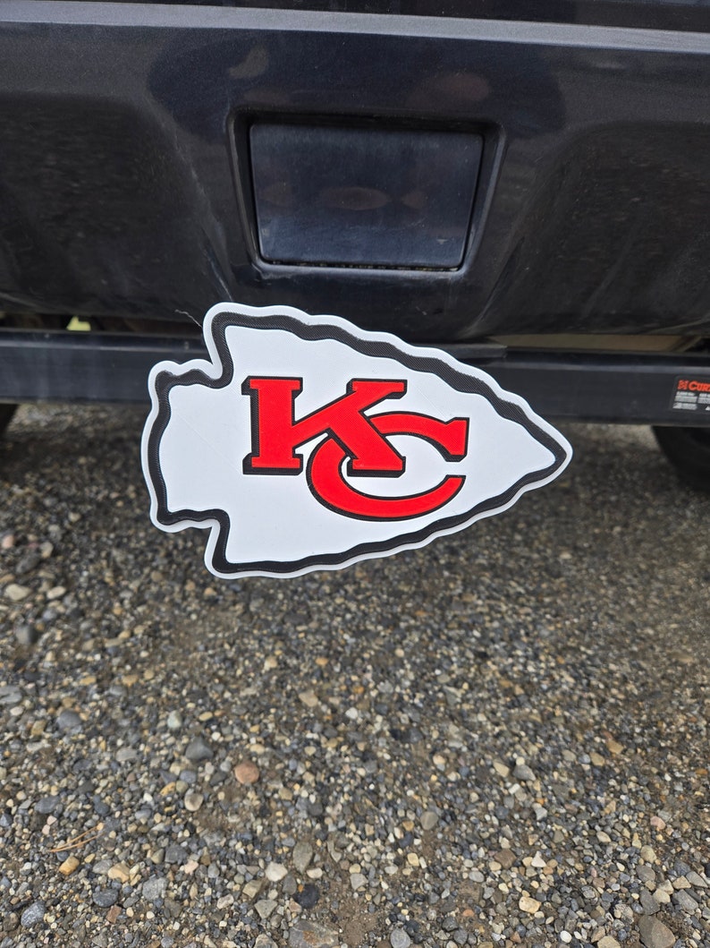 K&ouml;nnte beinhalten: Ein wei&szlig;er Anh&auml;ngerkupplungsschutz in Pfeilform mit dem Logo der Kansas City Chiefs in Wei&szlig; und Rot.