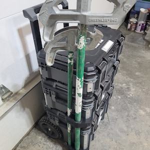 Może przedstawiać: Czarna skrzynia narzędziowa na kółkach z uchwytem. Dwa zielono-białe pręty są przymocowane do boku skrzyni. Skrzynia ma etykietę z napisem "GREENLEE 842". Na górze skrzyni znajdują się trzy srebrne giętarki do rur.