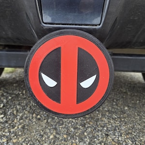 May include: A round, black and red trailer hitch cover featuring the Deadpool logo. The logo is a stylized face with white eyes and a red outline. The cover is attached to a black vehicle.