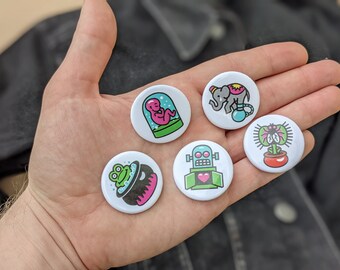Könnte beinhalten: Fünf runde weiße Anstecknadeln mit bunten Comic-Illustrationen. Die Nadeln zeigen einen Frosch in einem Kessel, einen Roboter mit einem Herzen, ein Baby in einem Glas, eine Pflanze mit einer Fliege und einen Elefanten auf einem Ball.