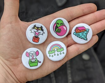 Könnte beinhalten: Fünf runde weiße Anstecknadeln mit bunten Comic-Illustrationen. Die Nadeln zeigen eine schwarze Katze, einen grünen Außerirdischen, der einen rosa Ball hält, ein Haus in einem Tornado, einen Regenbogen mit Regentropfen und einen weinenden Clown in einer Kiste.