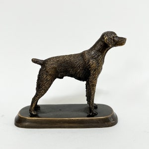 May include: A bronze sculpture of a dog standing on a base. The dog is brown and has a short, smooth coat. The sculpture is about 4 inches tall.