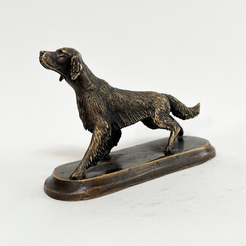 May include: A bronze sculpture of a dog, possibly a setter, standing on a wooden base. The dog is depicted in a hunting pose, with its head turned to the side and its tail raised.