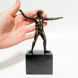 May include: Bronze sculpture of a nude male figure standing on a black marble base. The figure is posed with arms outstretched and has a beard.