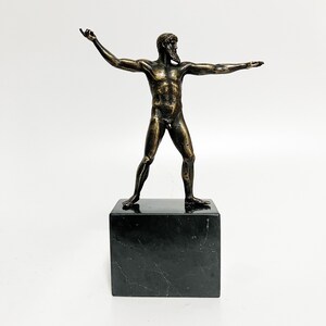 May include: Bronze sculpture of a nude male figure standing on a black marble base. The figure is posed with arms outstretched and has a beard.
