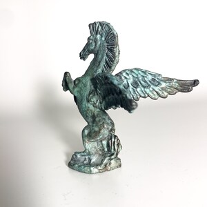 May include: A green bronze statue of a winged horse, or pegasus, with its wings spread out. The statue is standing on a base that looks like a wave.