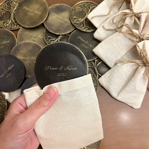 May include: A set of round, brass coasters featuring a tree of life design and the inscription "Pinar & Kerem 2022". The coasters are packaged in small, white fabric bags.