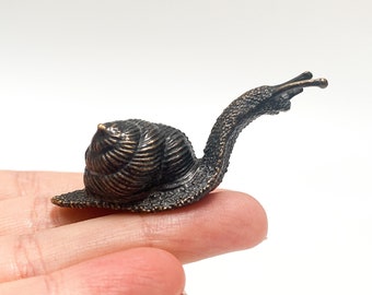 May include: A small bronze snail figurine with a detailed shell and a curled body. The snail is posed as if it is crawling forward.