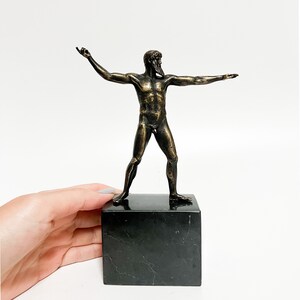 May include: Bronze sculpture of a nude male figure standing on a black marble base. The figure has a beard and is posed with arms outstretched.