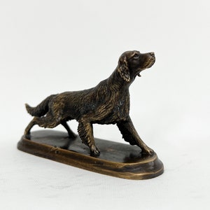 May include: A bronze sculpture of a dog, possibly a setter, standing on a small oval base. The dog is depicted in a running pose with its head turned to the side.