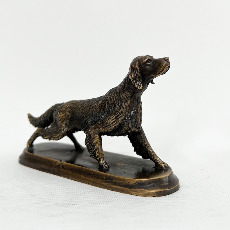 May include: A bronze sculpture of a dog, possibly a setter, standing on a small oval base. The dog is depicted in a running pose with its head turned to the side.