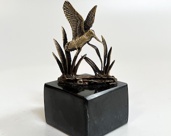 May include: A bronze sculpture of a bird in flight, perched on a black marble base. The bird is depicted with outstretched wings and a long beak, surrounded by stylized grass.