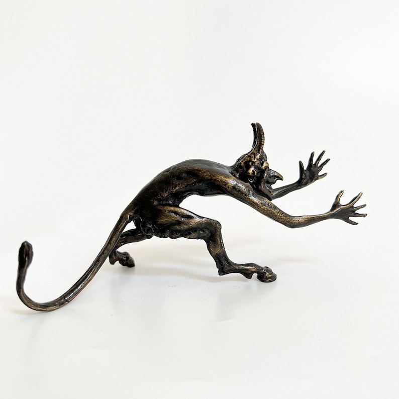 May include: A bronze sculpture of a small, horned demon with a long tail, crouching and reaching out with both hands. The demon has a fierce expression on its face.
