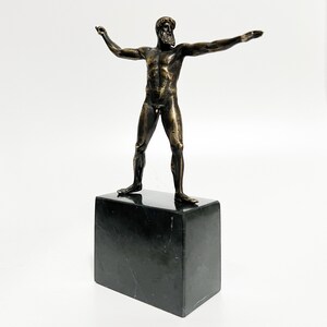 May include: Bronze sculpture of a nude male figure standing on a black marble base. The figure has a beard and is posed with arms outstretched.
