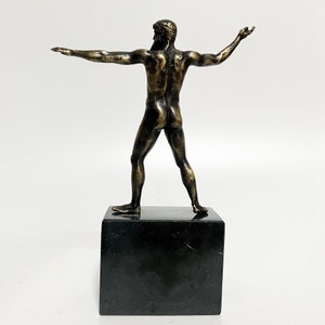 May include: Bronze sculpture of a nude male figure standing with arms outstretched. The sculpture is on a black marble base.