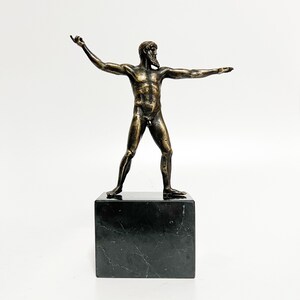 May include: Bronze sculpture of a nude male figure standing with arms outstretched on a black marble base.