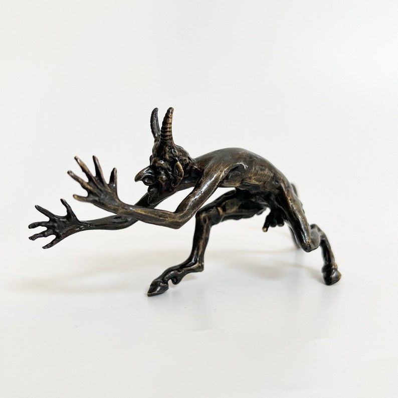 May include: A bronze sculpture of a horned demon with a hunched posture and outstretched arms. The demon has a long, pointed nose and a fierce expression.