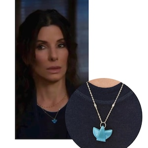 May include: A silver-toned chain necklace featuring a small, smooth, light blue pendant shaped like a stylized bird or angel with outstretched wings. The pendant hangs from a circular jump ring attached to a delicate metal link chain.