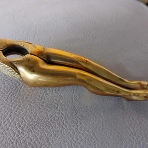 May include: A vintage brass nutcracker shaped like a reclining woman. The gold-coloured nutcracker has detailed legs, feet, and a head with a decorative element. This is a classic example of a figural nutcracker, often used for cracking nuts.