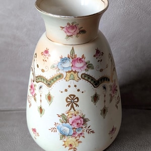May include: A cream-coloured ceramic vase featuring a flared rim with gold trim. The body is decorated with hand-painted floral clusters in pink, blue, and yellow, accented by intricate gold filigree, geometric patterns, and a central bow motif.