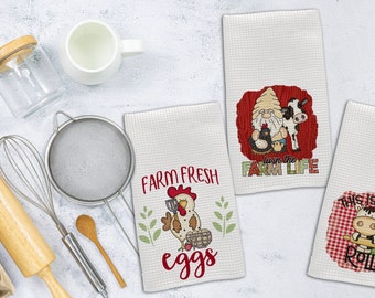 Könnte beinhalten: Drei weiße Küchentücher mit rot-weißem Karomuster. Die Handtücher zeigen Illustrationen und Texte zum Thema Bauernhof. Ein Handtuch sagt "Farm Fresh Eggs" mit einer Illustration eines roten Hahns. Ein anderes Handtuch sagt "Love the Farm Life" mit einer Illustration eines Zwergs und einer Kuh. Das dritte Handtuch sagt "This is How I Roll" mit einer Illustration einer Kuh.